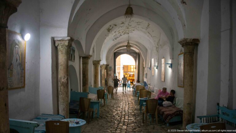Long vaulted medina passage with stone columns, cobbled paving, café tables, seated patrons, and pedestrians.