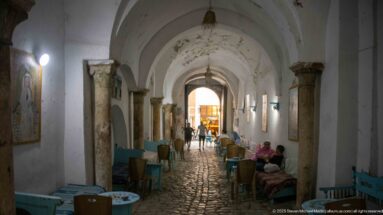 Long vaulted medina passage with stone columns, cobbled paving, café tables, seated patrons, and pedestrians.
