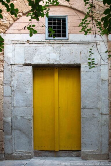Yellow double door set in a pale stone doorway beneath a small barred window, with vines crossing the wall.