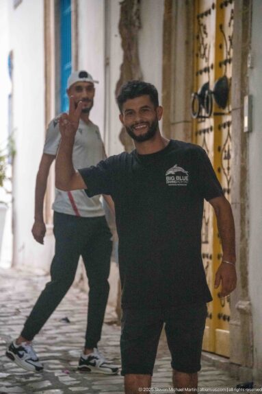 Smiling man raising his hand in a narrow medina lane, with another man behind him and a yellow doorway at right.