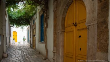 Narrow cobblestone lane with a tall yellow arched door at right and another yellow doorway in the distance.