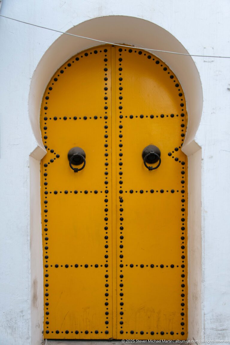 Rounded yellow double door covered with black metal studs and fitted with two dark knockers in a white arched recess.