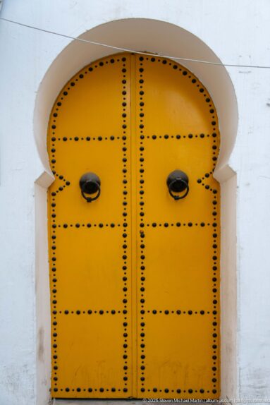 Rounded yellow double door covered with black metal studs and fitted with two dark knockers in a white arched recess.