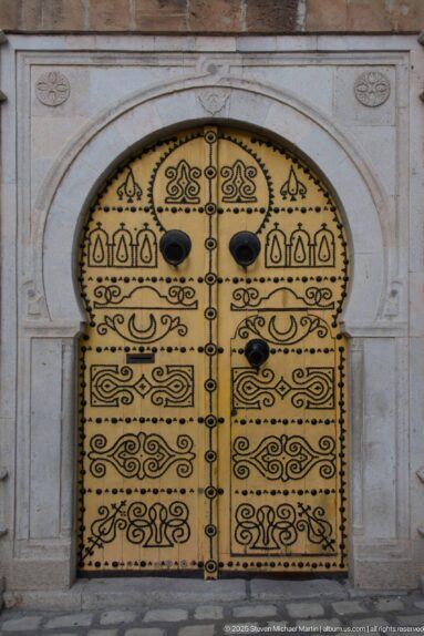 Ornate yellow arched double door covered with black scrollwork, geometric motifs, studs, and paired knockers.