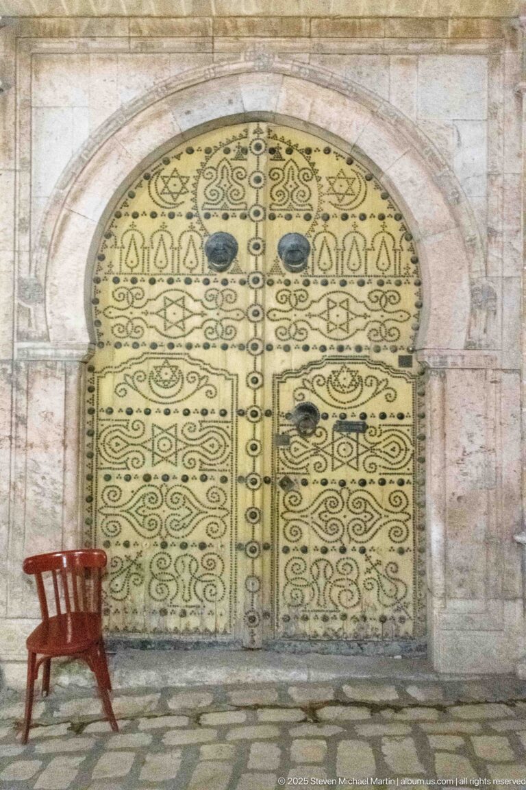 Ornate pale-yellow arched door with dark metal decoration and a red wooden chair on the cobbles at left.