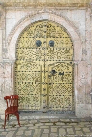 Ornate pale-yellow arched door with dark metal decoration and a red wooden chair on the cobbles at left.