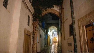 Nighttime cobblestone lane passing beneath several arches, with white walls and a bright opening in the distance.