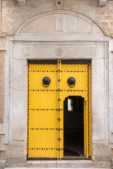 Bright yellow studded double door in a pale carved stone portal, with a small wicket door standing open.
