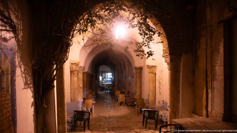Café tables and chairs arranged beneath a large masonry arch, with warm lights and more arches receding into the interior.