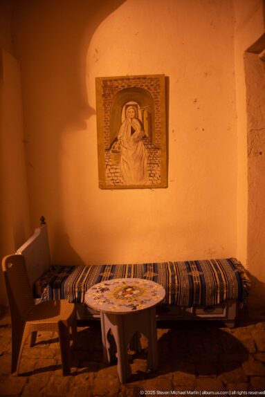 Striped bench, small round table, and chair against an ochre wall beneath a framed relief of a seated figure.
