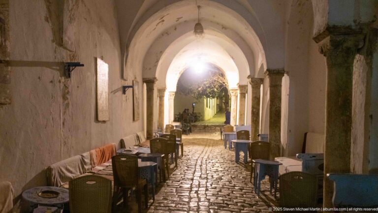 Café tables and chairs line a cobblestone passage beneath white vaults and stone columns, opening to a brighter lane beyond.
