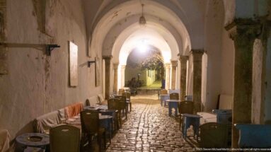 Café tables and chairs line a cobblestone passage beneath white vaults and stone columns, opening to a brighter lane beyond.