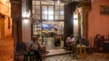 Two men seated at small tables beside a brightly lit café entrance framed by stone columns and an arched passage.