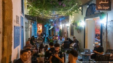 Crowded outdoor café filling a narrow lane beneath colored string lights, with blue doors, white walls, and flowering greenery.