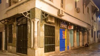 Corner façade with patterned tiles, dark decorative panels, pale columns, and a bright blue door beneath modern air-conditioning units.