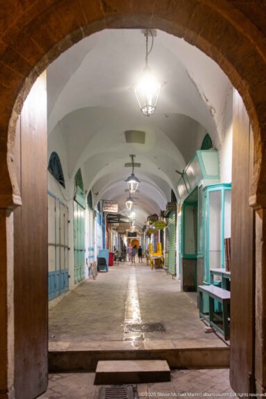 Long empty covered market aisle with pale blue shutters and turquoise counters, framed by a large masonry arch.