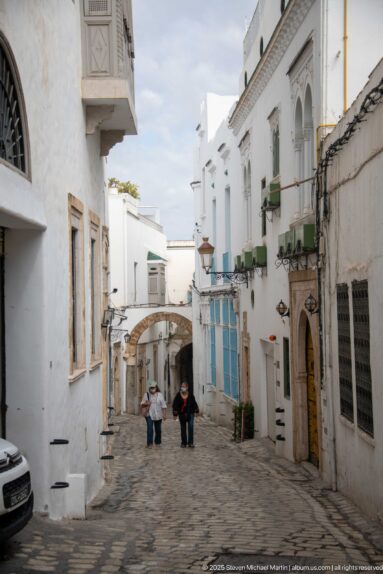 Sloping cobbled street between whitewashed medina houses, with two pedestrians walking toward a low arch in the distance.