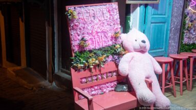 Large white teddy bear on a pink chair beside a floral neon display, turquoise doors, and pink café stools.