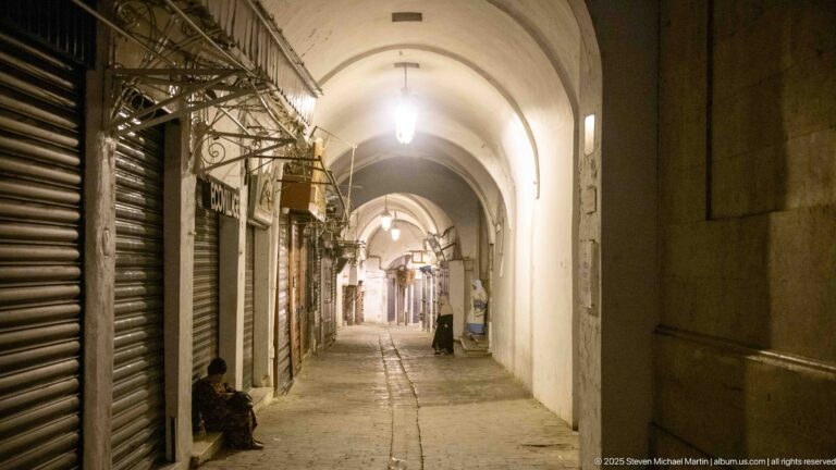 Narrow covered lane with closed metal shop shutters beneath repeated pointed white vaults and a few people along the sides.