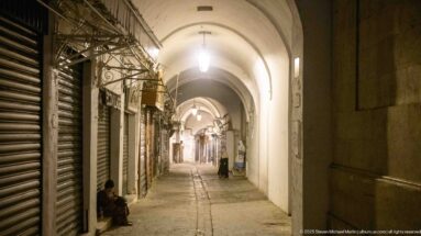 Narrow covered lane with closed metal shop shutters beneath repeated pointed white vaults and a few people along the sides.
