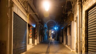 Empty nighttime commercial alley lined with closed metal shutters, bright lamps, tiled walls, and small awnings.