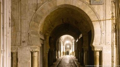 Deep carved stone arch with columns framing a narrow covered street of repeating arches and lamps.