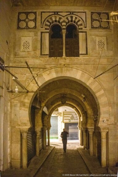 Broad stone arcade with thick columns and layered arches, paired windows above, and a lone pedestrian silhouetted at the far opening.