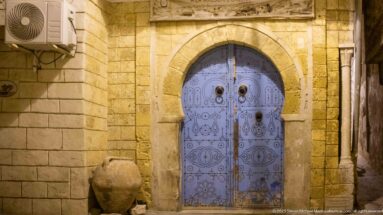 Ornate blue arched double door with symmetrical metal patterns and paired knockers, with a large ceramic jar standing at left.