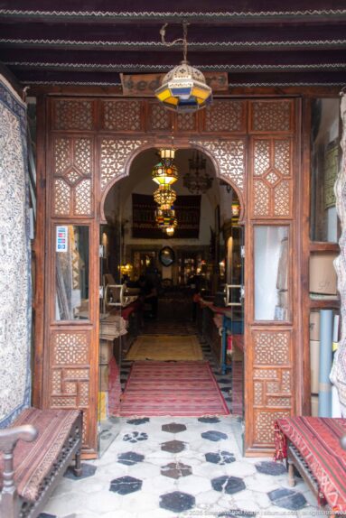 Carved wooden screen and arch opening into a furnished interior with hanging lamps, carpets, benches, and patterned flooring.