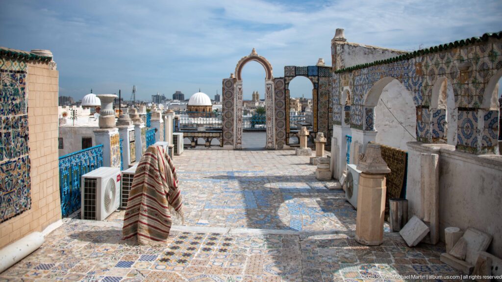 Multicolored tiled rooftop terrace with open arches and blue railings overlooking white domes and the Tunis skyline.