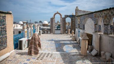 Multicolored tiled rooftop terrace with open arches and blue railings overlooking white domes and the Tunis skyline.
