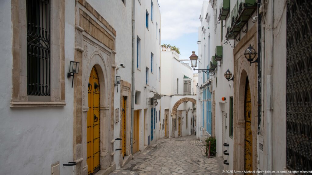 Descending medina lane with yellow doors, carved stone surrounds, wall lanterns, and a small arch at the far end.