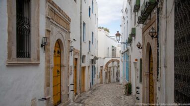 Descending medina lane with yellow doors, carved stone surrounds, wall lanterns, and a small arch at the far end.