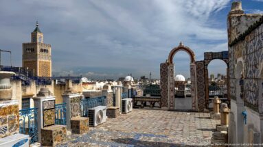 Tiled rooftop terrace with blue railings and arched screens overlooking white roofs, domes, and a prominent square minaret.