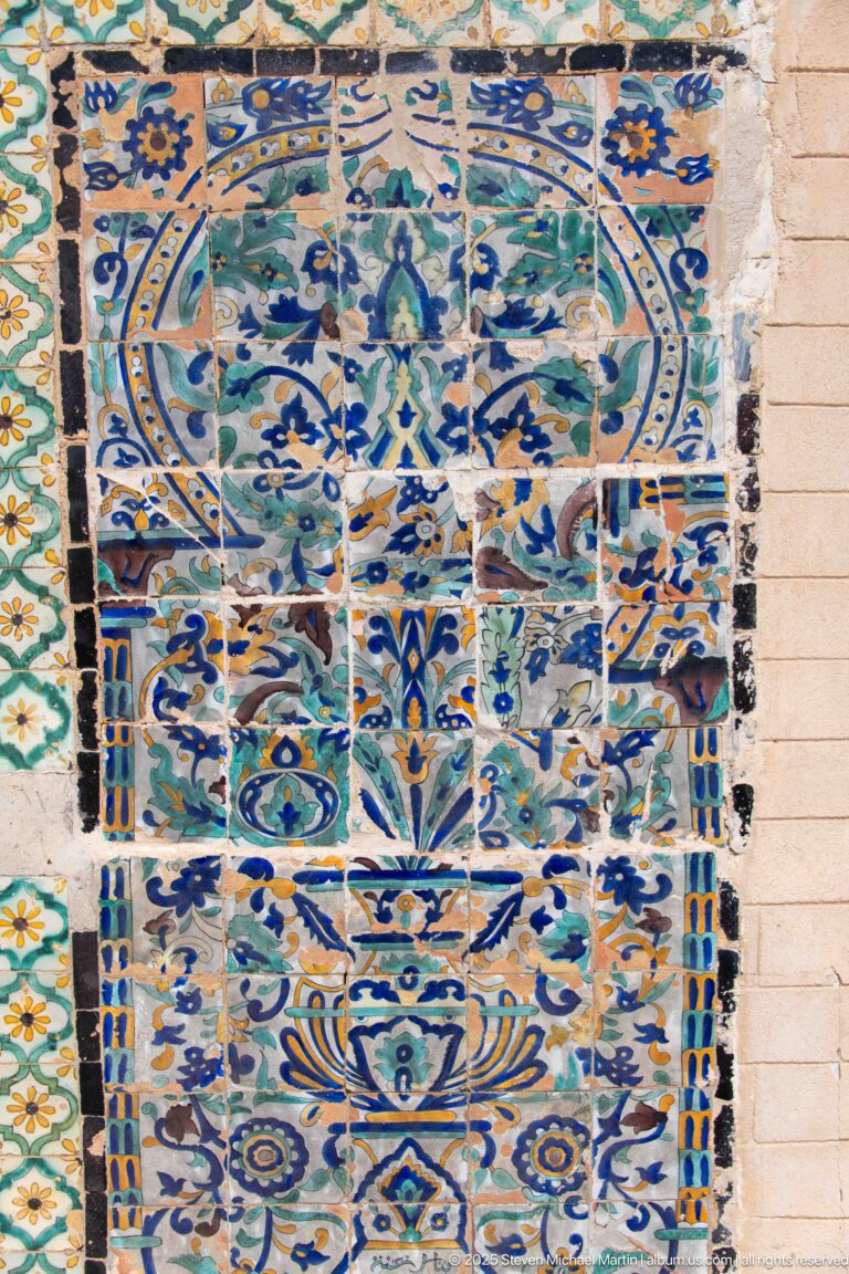 Large wall panel of blue, turquoise, green, and ochre ceramic tiles with floral and geometric motifs and visible weathering.