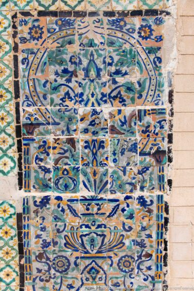 Large wall panel of blue, turquoise, green, and ochre ceramic tiles with floral and geometric motifs and visible weathering.