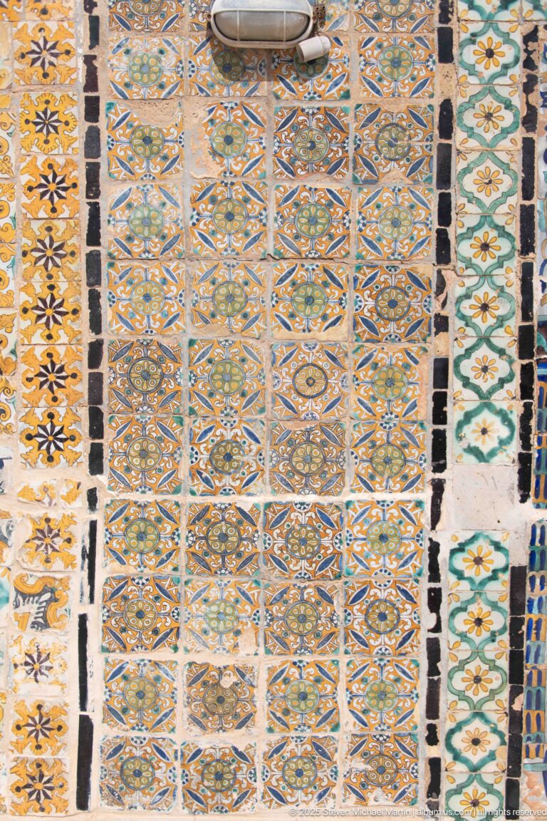 Wall covered with repeating geometric and floral ceramic tiles in blue, green, ochre, black, and white.