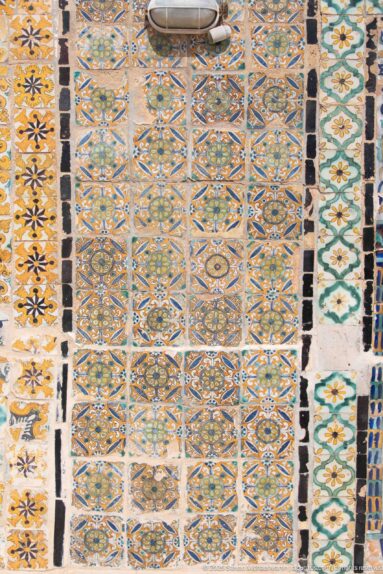 Wall covered with repeating geometric and floral ceramic tiles in blue, green, ochre, black, and white.