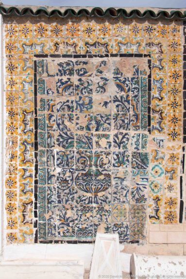 Weathered rectangular ceramic tile panel in blue, turquoise, ochre, and white, framed by multiple patterned borders.