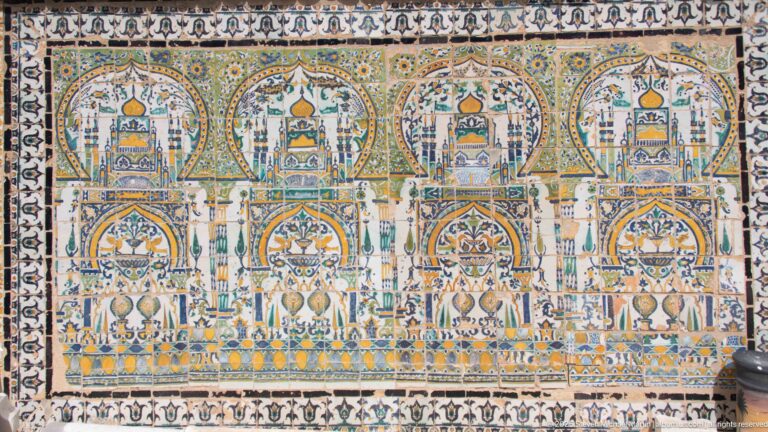 Large symmetrical tile mural with repeated pointed arches, towers, floral motifs, and geometric borders in blue, green, and ochre.