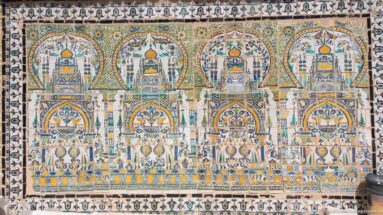 Large symmetrical tile mural with repeated pointed arches, towers, floral motifs, and geometric borders in blue, green, and ochre.