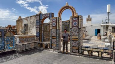 Visitor standing beneath a tall tiled rooftop archway with smaller arches, railings, white roofs, and a tower in the background.