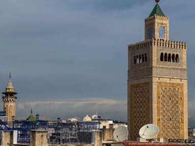 Square minaret with patterned panels, arched openings, and a green pyramidal roof rising above rooftops and satellite dishes.
