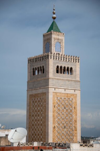 Square minaret with broad geometric panels, arched upper gallery, green-tiled roof, and finial against a blue-gray sky.