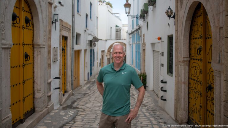 Man in a green shirt standing on a narrow cobbled medina lane between ornate yellow doors and white façades.