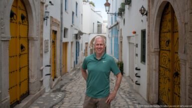 Man in a green shirt standing on a narrow cobbled medina lane between ornate yellow doors and white façades.
