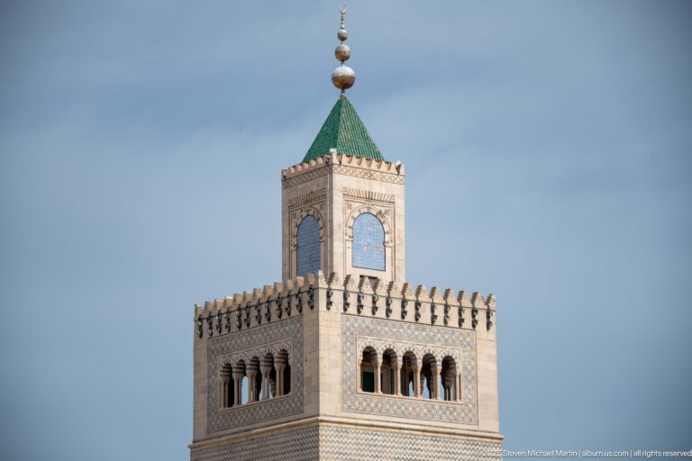 Close view of a square minaret’s arched gallery, patterned masonry, green pyramidal roof, and stacked finial.