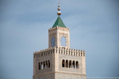 Close view of a square minaret’s arched gallery, patterned masonry, green pyramidal roof, and stacked finial.