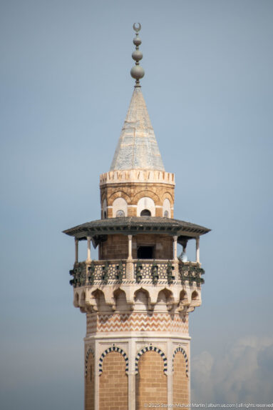 Ornate tower with projecting balcony, small arches, patterned bands, and a tapered pale roof topped by a finial.