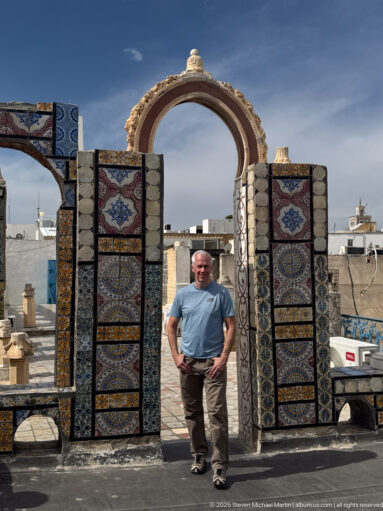 Visitor standing beneath a large multicolored tiled rooftop arch with city rooftops visible through and around the opening.